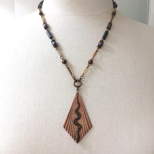 May include: A copper pendant necklace with a diamond-shaped pendant featuring a textured design and a dark snake detail. The necklace has a beaded chain with dark and gold-toned beads.