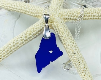 May include: A cobalt blue pendant necklace in the shape of the state of Maine, with a heart-shaped cutout. The pendant hangs from a delicate silver chain. The necklace is displayed on a white starfish.