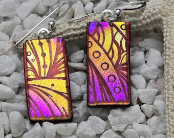 May include: A pair of rectangular glass earrings with a red, yellow, and purple iridescent finish. The earrings feature a geometric design with swirls and circles. The earrings are attached to silver hooks.