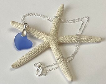 May include: A blue heart-shaped pendant necklace with a silver chain. The pendant is a matte finish and is suspended from a silver bail. The necklace is displayed on a white starfish.