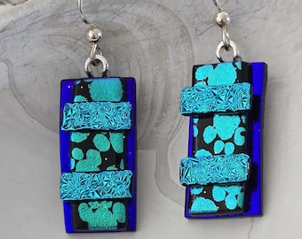 May include: A pair of blue and green glass earrings with silver hooks. The earrings are made of layered glass with a textured surface.