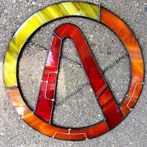 May include: A stained glass emblem featuring a stylized 'A' within a circle. The design incorporates vibrant colors, including yellow, orange, and red, with a textured appearance. The glass is framed with a dark border.