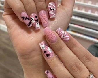 May include: A hand with long, coffin-shaped, pink acrylic nails. The nails feature Halloween-themed designs, including stitches, ghosts, spiderwebs, and mummy-like figures. The designs are in black and white, with a pink base.