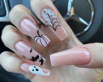 May include: Close-up of a hand with long, nude-colored acrylic nails decorated with Halloween-themed designs. The designs include a spiderweb, a pumpkin, a bat, and a ghost.