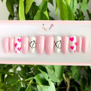 Puede incluir: Conjunto de diez u&ntilde;as postizas rosas y blancas con diferentes dise&ntilde;os. Algunas u&ntilde;as tienen corazones rosas, otras tienen las letras "XO" en negro.