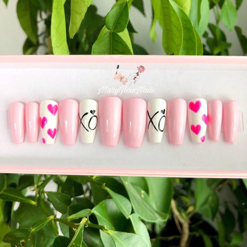 Puede incluir: Conjunto de diez u&ntilde;as postizas rosas y blancas con diferentes dise&ntilde;os. Algunas u&ntilde;as tienen corazones rosas, otras tienen las letras "XO" en negro.