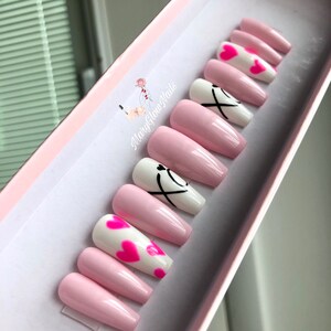 Puede incluir: U&ntilde;as postizas largas de color rosa y blanco con un dise&ntilde;o de corazones y las letras "XO" en negro.