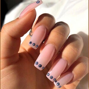 May include: Close-up of a hand with long, light pink acrylic nails. Each nail is decorated with a row of small, blue evil eye charms.