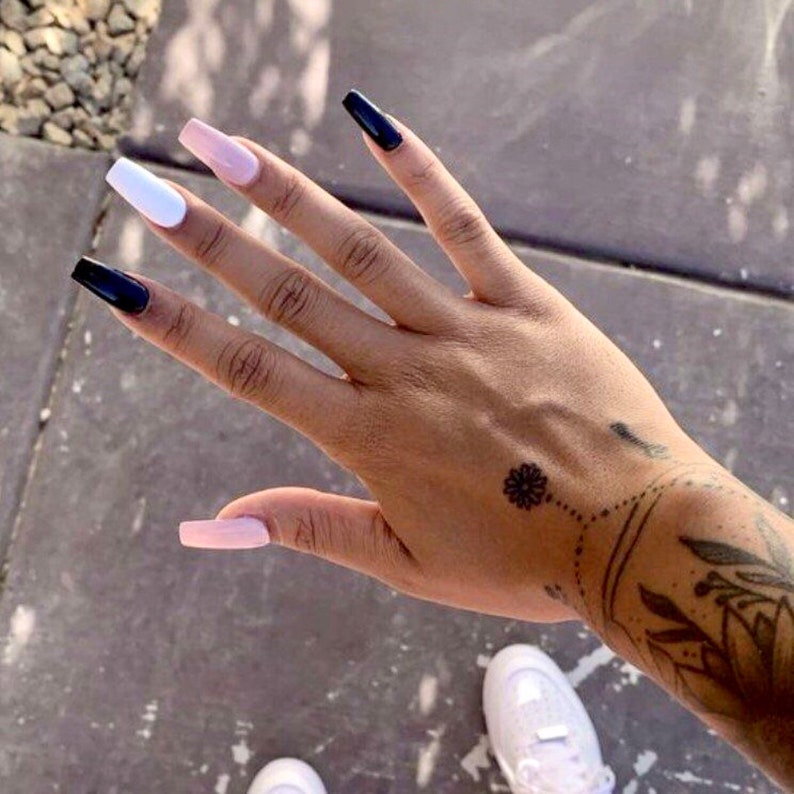 May include: Close-up of a hand with long, square-shaped nails painted in black, white, and pink. The hand is resting on a gray surface. The person has a small flower tattoo on their wrist.