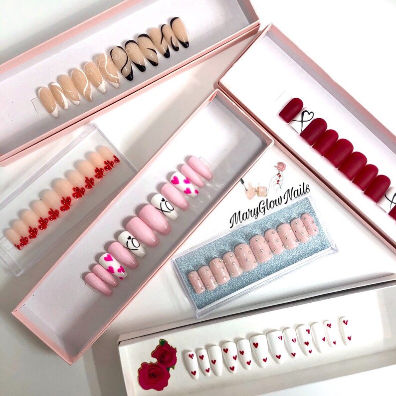 Puede incluir: Cinco cajas de u&ntilde;as postizas con diferentes dise&ntilde;os. Los dise&ntilde;os incluyen corazones, remolinos y pedrer&iacute;a. Las cajas son rosas y blancas con el texto "MaryGlow Nails" en una de las cajas.