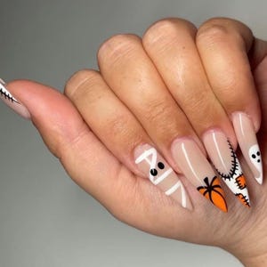 May include: Long almond-shaped artificial nails featuring a nude base with various Halloween-themed designs including a white mummy wrap, an orange pumpkin, a black stitched patch, and a white ghost with black eyes on the tips.