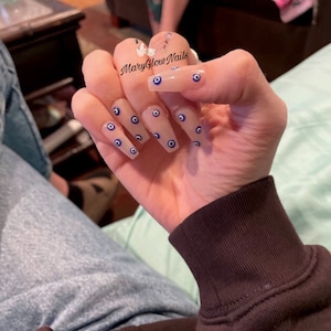 May include: Long, nude-colored fake nails with a blue and white evil eye design. The nails are decorated with multiple evil eye charms.