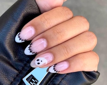 Puede incluir: Primer plano de una mano con uñas en forma de almendra pintadas con un esmalte rosa claro. Las uñas tienen una punta blanca con un diseño cosido negro. Una uña presenta un fantasma blanco con ojos negros.