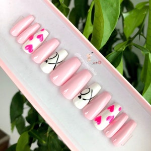 Puede incluir: U&ntilde;as postizas en forma de ata&uacute;d de color rosa y blanco con corazones rosas y dise&ntilde;os negros de "XO".