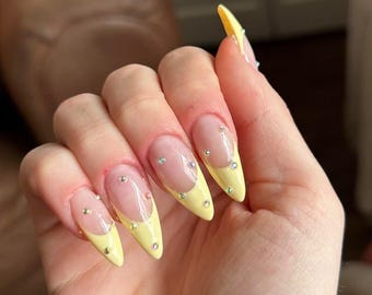 Puede incluir: Una mano con uñas largas en forma de almendra. Las uñas presentan una base transparente, una punta francesa amarilla y pequeños brillantes. Las uñas están bien cuidadas y tienen un acabado brillante.