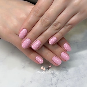 May include: A set of manicured nails painted light pink with white polka dots. The nails are oval-shaped and the design is consistent across all nails. The image includes the text "Mary Glow Nails".