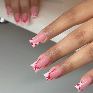 May include: Pink acrylic nails with a French tip design. Some nails feature red heart accents, while others have red bow designs. The nails are square-shaped and have a glossy finish.