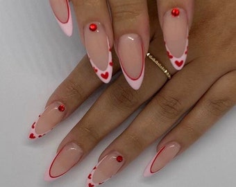 May include: Close-up of long, almond-shaped nails with a nude base and a white outline. The outline is decorated with small red hearts. Some nails have a single red rhinestone.