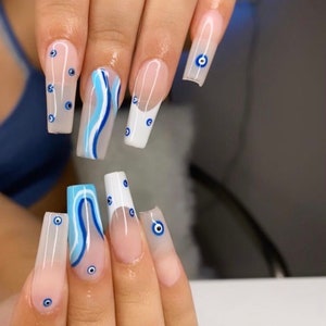 May include: Close-up of a hand with long, white, and blue painted nails. The nails are decorated with a blue and white wave pattern and blue evil eye charms.