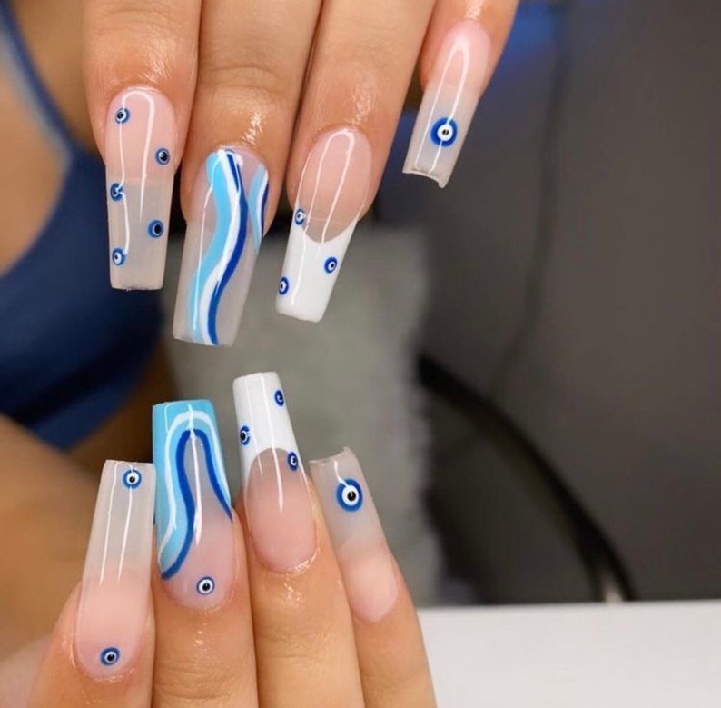 May include: Close-up of a hand with long, white, and blue painted nails. The nails are decorated with a blue and white wave pattern and blue evil eye charms.