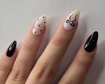 May include: A hand with almond-shaped nails. Two nails are painted solid black, one is nude with a black bow design, and another is nude with black polka dots. The nails are long and well-manicured.