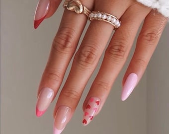 May include: Close-up of a hand with long, almond-shaped nails painted in a light pink color. Some nails have a red tip, while others have small red hearts scattered across the nail. The hand is wearing two gold rings.