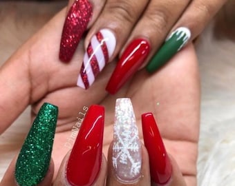 May include: A hand with long, coffin-shaped acrylic nails. The nails feature a variety of festive designs, including red glitter, candy cane stripes, solid red, a snowflake, and green glitter. The nails are painted with a glossy finish.