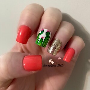 May include: Close-up of a hand with short, square-shaped nails. Four nails are painted a bright coral color. One nail is painted white with a green cactus design and pink flowers. Another nail is covered in gold glitter. The image includes the text "MaryGlowNails".