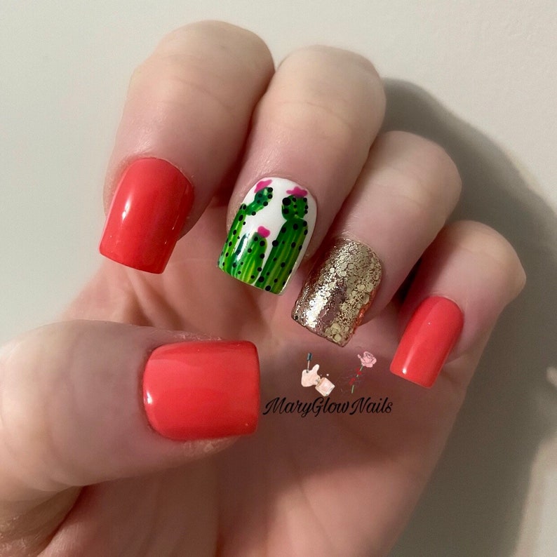 May include: Close-up of a hand with short, square-shaped nails. Four nails are painted a bright coral color. One nail is painted white with a green cactus design and pink flowers. Another nail is covered in gold glitter. The image includes the text "MaryGlowNails".