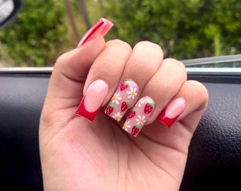 May include: Close-up of a hand with long, square-shaped nails. The nails are painted with a light pink polish and have a red tip. The nails are decorated with a design of strawberries and white flowers.