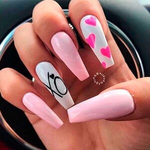 Puede incluir: U&ntilde;as acr&iacute;licas largas en forma de almendra, de color rosa y blanco, con un dise&ntilde;o de corazones rosas en una u&ntilde;a y las letras XO con un peque&ntilde;o coraz&oacute;n en otra u&ntilde;a.
