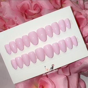 May include: A set of light pink press-on nails in various sizes, arranged on a white card. The nails have a glossy finish and an oval shape. The card is surrounded by pink fabric and features the text "MaryGlowNails".