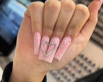 Puede incluir: Uñas acrílicas largas y cuadradas de color rosa con pedrería y un diseño en forma de corazón en el dedo corazón.