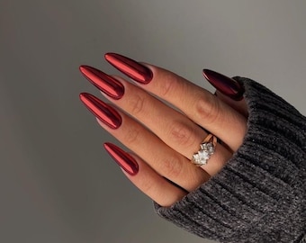 May include: Close-up of a hand with long, red, stiletto-shaped nails. The nails are painted with a shiny, metallic red polish. The hand is wearing a gold ring with a large, clear stone.