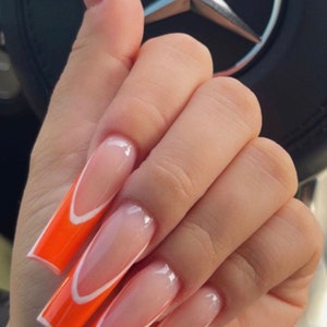 May include: Close-up of a hand with long, orange and white, almond-shaped nails. The nails have a white tip with a curved edge and a solid orange base.