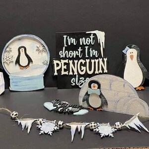 May include: A set of penguin-themed wooden decorations, including a snow globe, a sign that reads "I'm not short I'm penguin size", and a penguin figurine. The decorations are painted in black, white, and blue.