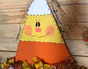 May include: A wooden candy corn shaped door hanger with a painted face and a bow made of raffia. The candy corn is decorated with orange, yellow, and white paint. The bottom of the door hanger is decorated with a bow made of orange, yellow, and white plaid ribbon.