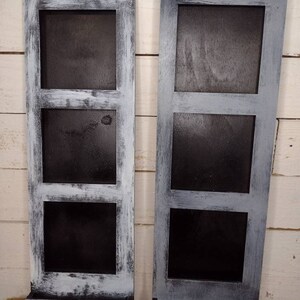 May include: Two vertical wooden picture frames with three rectangular openings each. The frames are painted white with a distressed finish, giving them a rustic look. The frames are standing on a wooden surface.