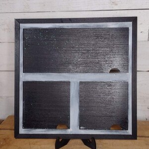 May include: A black framed chalkboard with a white border and three cut-out sections. The chalkboard is standing on a wooden surface.