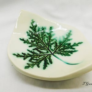 May include: A white ceramic dish shaped like a leaf with a green leaf design painted on it. The dish is glazed and has a glossy finish. The artist's signature, "J. Gant", is visible in the lower right corner.