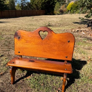 May include: A small wooden bench with a warm brown finish, featuring a backrest with a cutout heart shape in the center. The bench is constructed with visible dark metal fasteners at the corners of the backrest and the seat, supported by four legs.