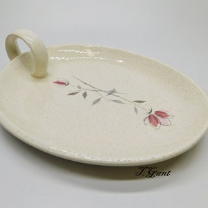 May include: A white oval serving dish with a handle. The dish has a speckled glaze and a floral design with pink flowers and gray stems.