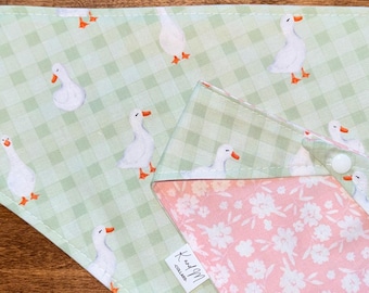Puede incluir: Un pañuelo triangular para mascotas con un patrón de cuadros vichy en verde claro y blanco, decorado con un estampado de ocas blancas con picos y patas naranjas. El reverso muestra una tela rosa suave con flores blancas y una etiqueta.