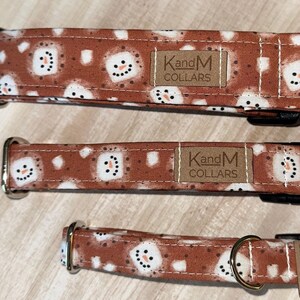 Op de afbeelding: Drie bruine hondenriemen met een sneeuwpopmotief. De riemen hebben een bruin lederen label met de tekst "KandM Collars".