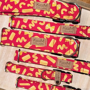 Peut inclure: Plusieurs colliers rouges pour chiens ornés de motifs de pâtes jaunes. Chaque collier est doté d'une boucle métallique et d'une étiquette en cuir avec le texte "KandM COLLARS". Les colliers sont présentés sur un plateau en bois.