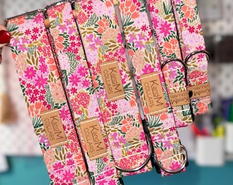 Puede incluir: Varios collares para perros con estampado floral en tonos rosa, naranja y verde. Cada collar tiene una hebilla metálica y una etiqueta de cuero con el texto "Kang M Collars". Los collares se muestran sobre un fondo turquesa.