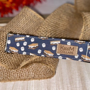 May include: A blue dog collar with a pattern of brown graham crackers, white marshmallows, and chocolate. The collar has a silver buckle and a brown leather tag that says "KandM Collars".