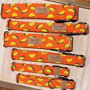 Può includere: Un assortimento di collari per cani arancioni con un motivo ripetuto di tacos gialli. Ogni collare ha una fibbia in metallo e una piccola etichetta con la scritta "Kan&M Collars". I collari sono disposti su un vassoio di legno.
