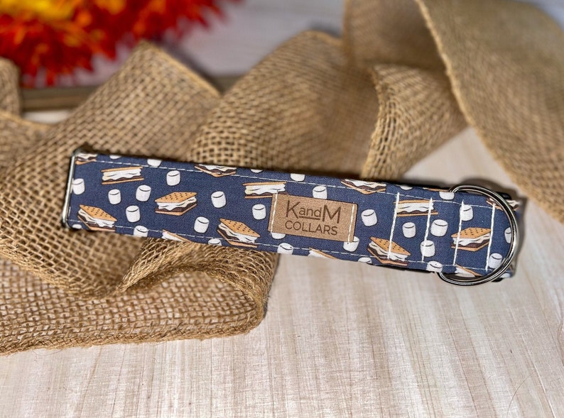 May include: A blue dog collar with a pattern of brown graham crackers, white marshmallows, and chocolate. The collar has a silver buckle and a brown leather tag that says "KandM Collars".