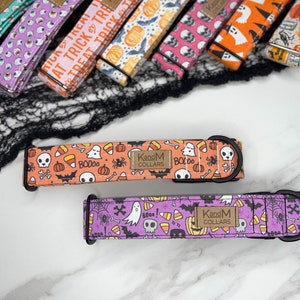 May include: Two Halloween-themed dog collars with orange and purple backgrounds. The collars feature a variety of spooky designs, including ghosts, skulls, bats, candy corn, and pumpkins. The collars are made by KandM Collars.